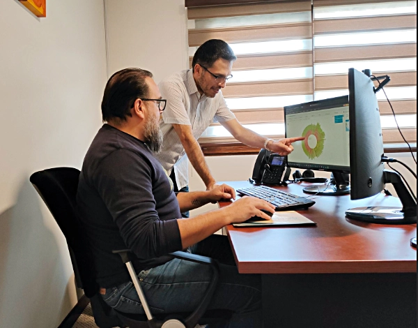 Senior ETTE technician pointing out a pie chart graphic in a computer monitor to a colleage sitting at his desktop.